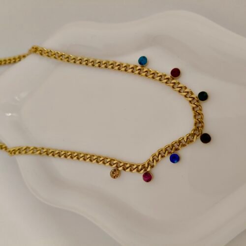 Χρυσή αλυσίδα τύπου "cuban link" διακοσμημένη με μικρές, πολύχρωμες κρεμαστές πέτρες σε διάφορα χρώματα, τοποθετημένη σε λευκή κεραμική βάση.