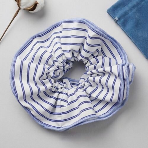 Λευκό υφασμάτινο scrunchie με λεπτές μπλε ρίγες και γαλάζιο ρέλι στο τελείωμα. Δίπλα του διακρίνονται ένα κλαδί βαμβακιού και ένα μπλε βελούδινο πουγκάκι.