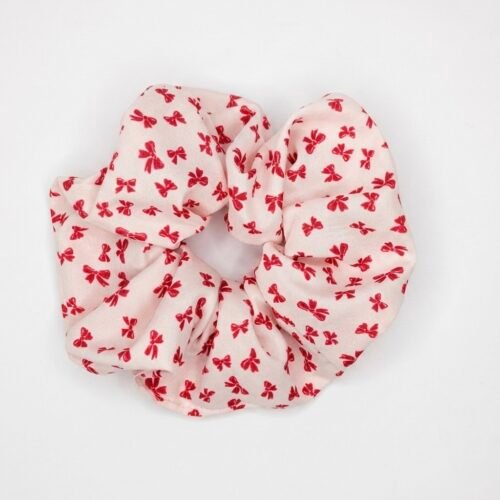 Υφασμάτινο scrunchie (λαστιχάκι μαλλιών) σε απαλή, ανοιχτή απόχρωση με μοτίβο από μικρούς κόκκινους φιόγκους, φωτογραφημένο σε καθαρό λευκό φόντο.